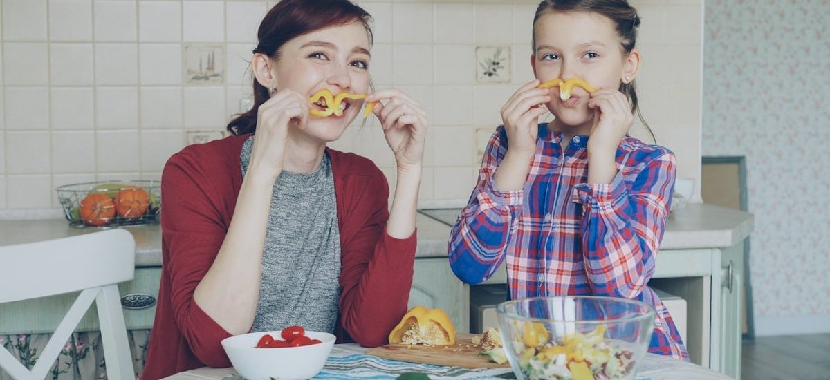 Joyful mother and cute cheerful daughter have fun grimacing silly with vegetables and looking into camera while cooking in the kitchen at home. Family, cook, and people concept