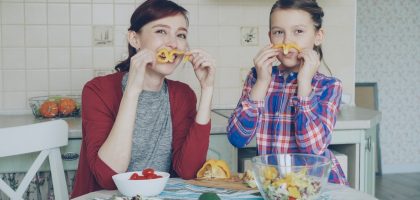 Joyful mother and cute cheerful daughter have fun grimacing silly with vegetables and looking into camera while cooking in the kitchen at home. Family, cook, and people concept