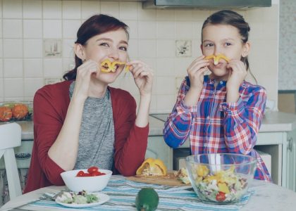 Joyful mother and cute cheerful daughter have fun grimacing silly with vegetables and looking into camera while cooking in the kitchen at home. Family, cook, and people concept