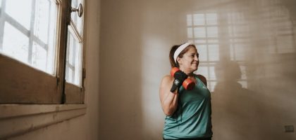 Senior woman in a brightly lit room exercises with dumbbells, promoting fitness at home.